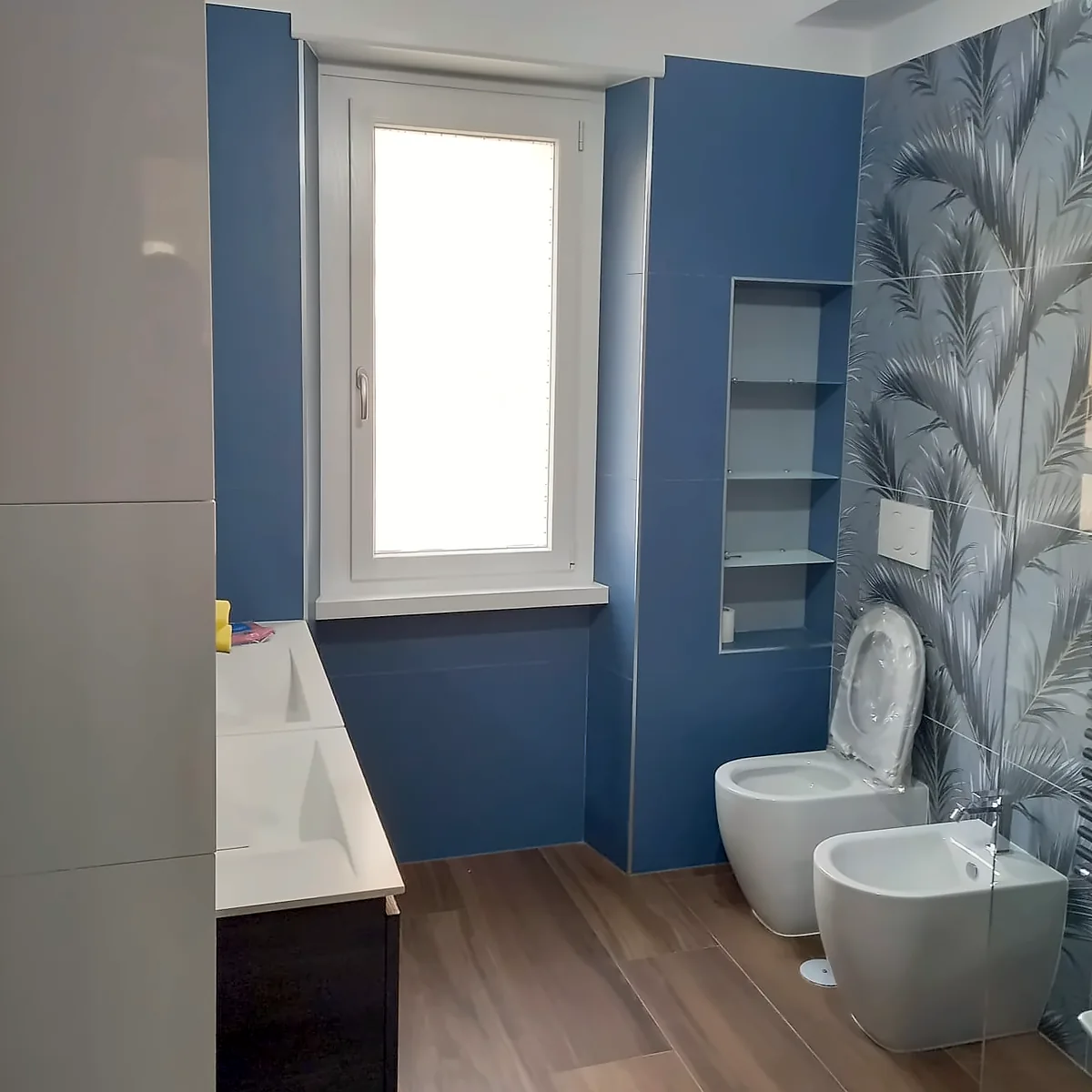 Bagno ristrutturato a Roma con pareti blu e carta da parati a foglie Bagno ristrutturato da Edil Dima a Roma: pareti blu, carta da parati a foglie nella zona sanitari, nicchia a giorno e pavimento effetto legno